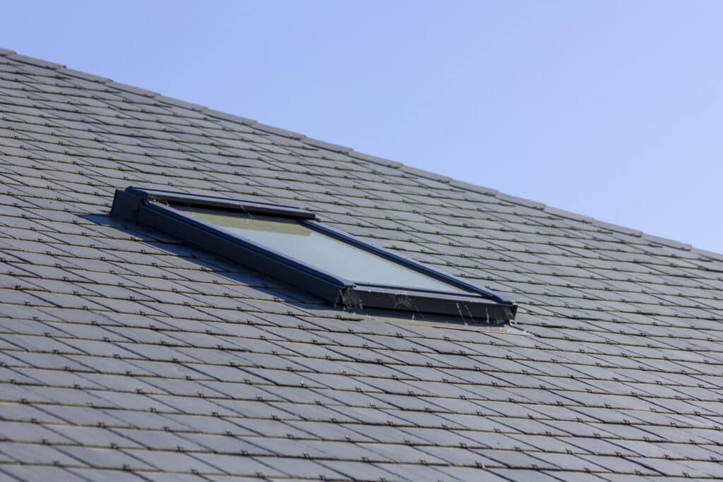 Toiture Ardoise avec Fenêtre de Toit : Détail Clair Fenêtre de toit Velux noire encastrée dans une toiture en ardoises grises sous un ciel bleu clair.