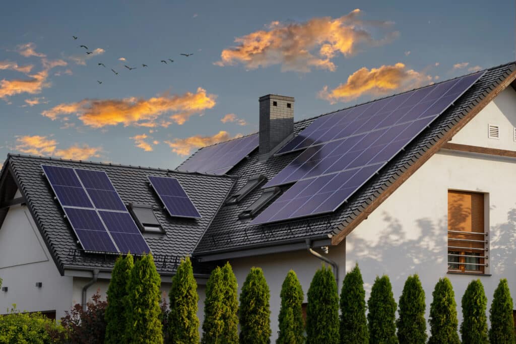 Panneaux solaires sur maison au coucher du soleil et oiseaux Maison moderne avec panneaux solaires sur le toit foncé, au coucher du soleil, des oiseaux volent au loin.