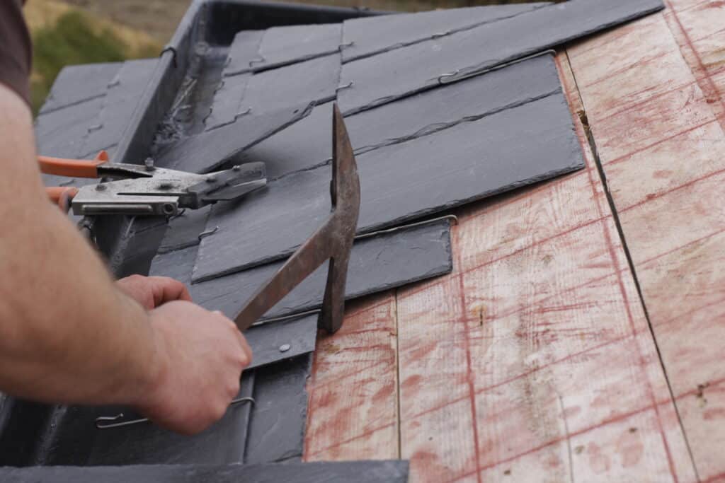 Pose d'ardoises de toiture : outils et fixation Couvreur utilisant un outil pour fixer des ardoises noires sur un sous-plancher en bois clair, vue rapprochée des mains.